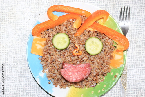 Breakfast for a child is decorated in the form of a funny face in a plate. Buckwheat, pepper, cucumber, cheese, sausage are good for the body