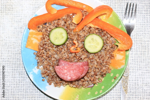 Breakfast for a child is decorated in the form of a funny face in a plate. Buckwheat, pepper, cucumber, cheese, sausage are good for the body