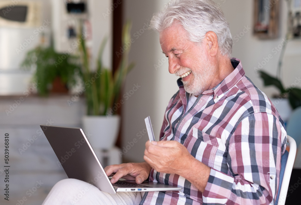 Elderly bearded man browsing by laptop enjoying shopping online. Joyful and smiling beautiful senior man at home being in great mood using credit card to purchase Elderly bearded man browsing by laptop enjoying shopping online. Joyful and smiling beautiful senior man at home being in great mood using credit card to purchase