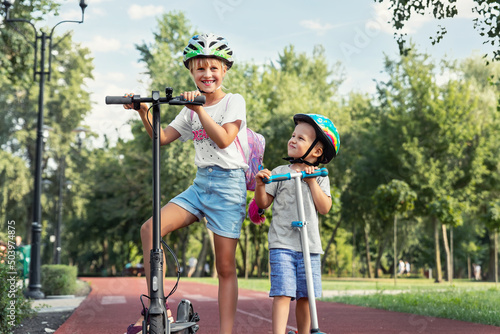 Portrait of two cute blond little caucasian sibling kids girl wear safety helmet enjoy having fun riding electric scooter city street park outdoors sunny day. Healthy sport children activity outside