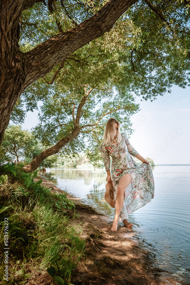 Fototapeta premium A young 30 year old woman in a long dress and barefoot walks along the beach lake. Beautiful landscape. Feminine style of a European girl in sunglasses and straw hat. Slim body. Natural environment