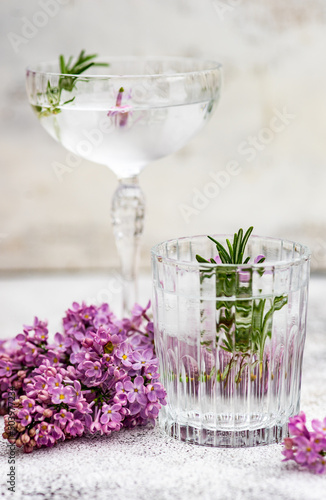 Glass of ice water and cocktail in a champagne coupe with a sprig of rosemary and lilac flowers photo