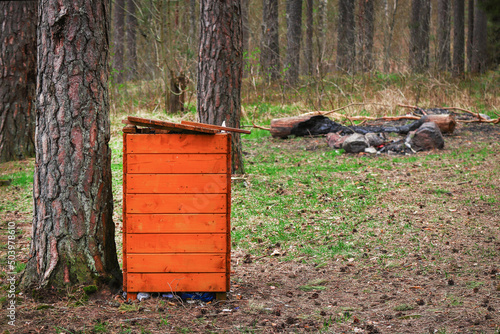 Wooden brigh orange trash bin in forest resting spot between tree trunks on green grass in spring