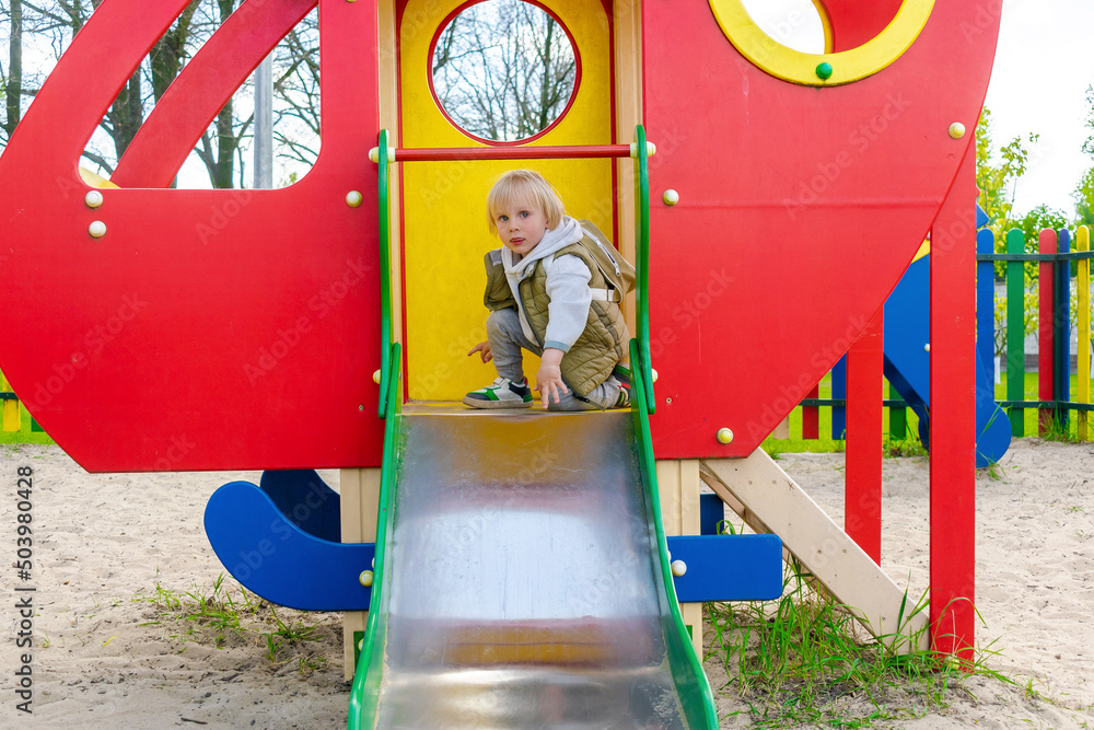 Cute funny smiling blonde little young toddler kid child boy going down slide in playground.Children physical,emotional development and childhood daycare, kindergarten concept
