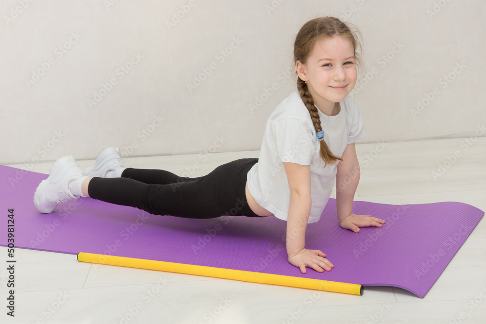 cute little girl stands in a plank on a mat, a gymnastic stick lies nearby, the child smiles cute little girl stands in a plank on a mat, a gymnastic stick lies nearby, the child smiles