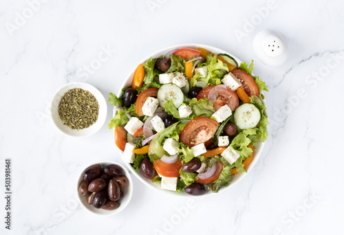 Greek salad of fresh vegetables - cucumbers, tomatoes, bell peppers, onions, feta cheese and olives on a white plate in a white bowl, top view.