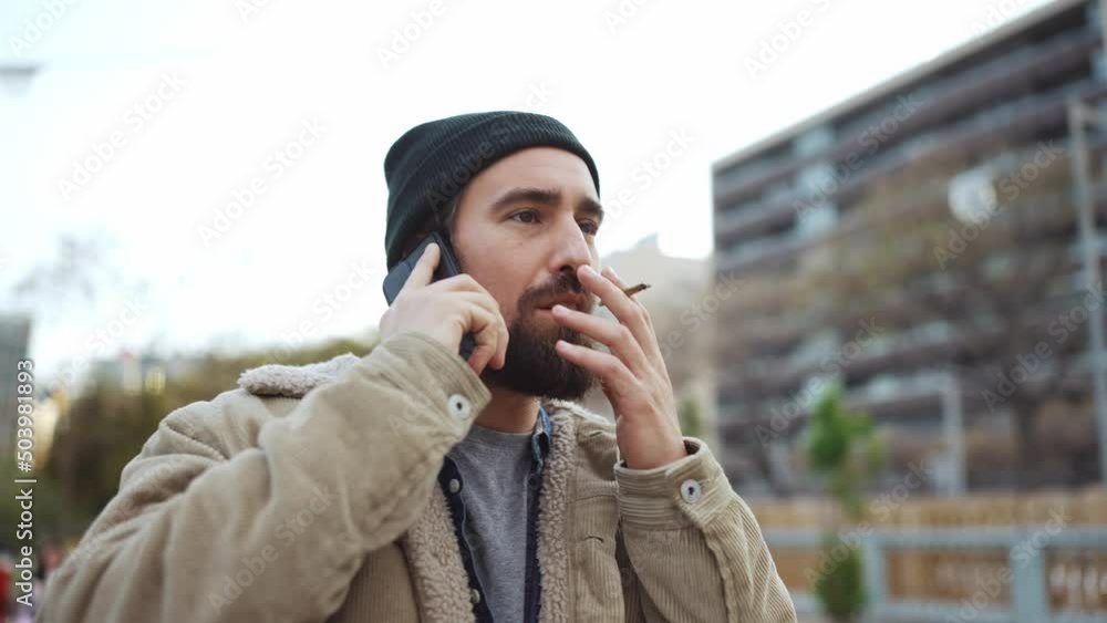 Smiling bearded man wearing hat talking by phone and smoking outdoors Smiling bearded man wearing hat talking by phone and smoking outdoors