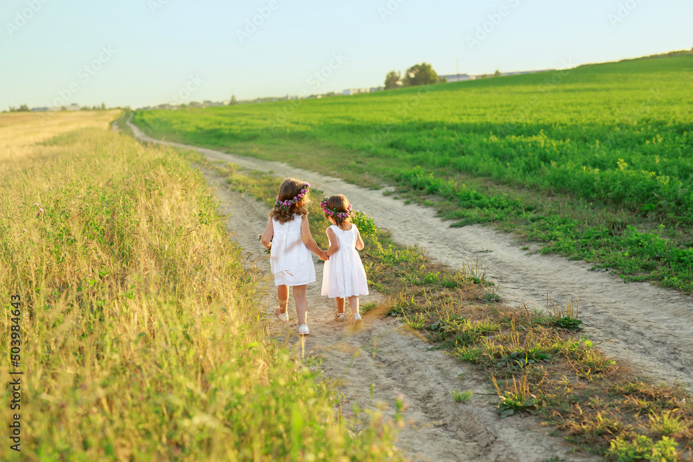 Children run hand in hand in the fresh air among fields and meadows on a narrow sandy path. Girls in dresses and with wreaths of flowers in their hair at sunset in nature Children run hand in hand in the fresh air among fields and meadows on a narrow sandy path. Girls in dresses and with wreaths of flowers in their hair at sunset in nature