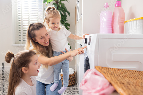 Obraz na płótnie Woman spends time with young children in laundry room, bathroom, performs household duties during maternity leave, takes care of daughters, shows girls how to operate washing machine