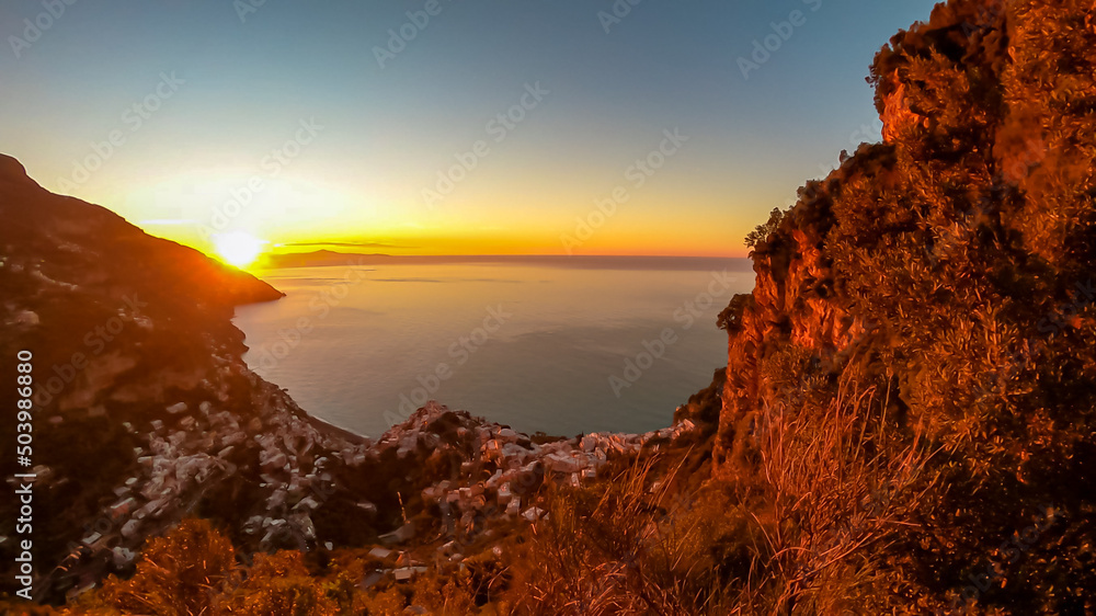 Panoramic sunrise view from hiking trail Path of the Gods between Positano and Praiano at Amalfi Coast, Campania, Italy, Europe. Calm water surface reflecting sun ray at Tyrrhenian Mediterranean Sea