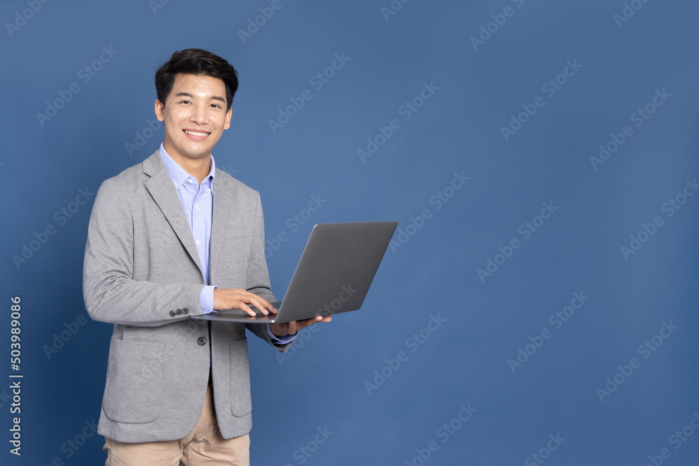 Young Asian business man using laptop computer and looking at camera isolated on blue background