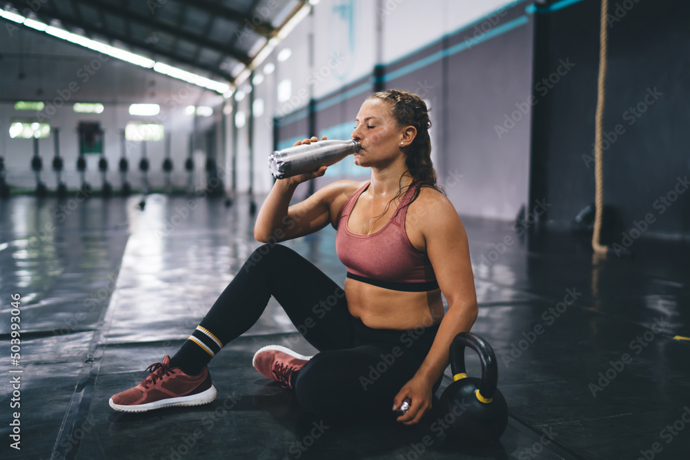 Side view of Caucasian female in activewear drinking water from bottle refreshing during sportive break in gym studio, fit girl athlete recreating during cardio workout taste protein cocktail indoors