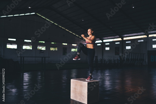 Young crossfit woman training at box jump exercising in sportive gym studio, determined Caucasian female athlete in tracksuit slimming during cardio practice and physical recreation indoors