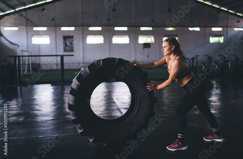Motivated female athlete in sportive tracksuit working out with tire - weightlifting in gym studio during slimming time, determined fit girl checking body strength during training practice indoors