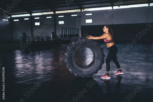 Cheerful woman 20 years old working out with training equipment during time in sport hall doing cardio exercises, smiling female athlete with wheel tire weightlifting in gym studio interior