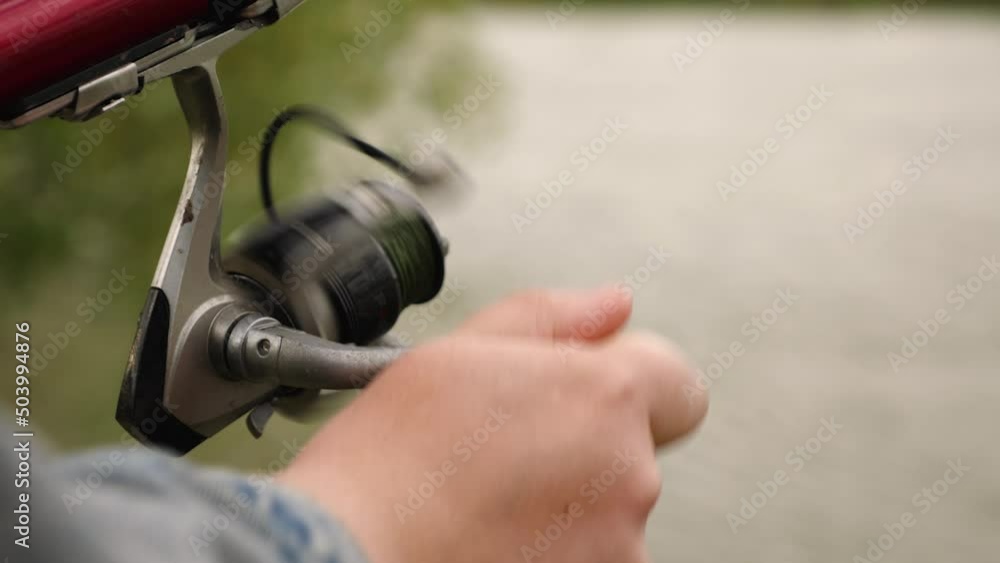 Close-up of a woman's hand spinning a fishing rod reel against a river background. Slow motion. The green fishing line is reeled in. The concept of recreation and hobbies in nature Close-up of a woman's hand spinning a fishing rod reel against a river background. Slow motion. The green fishing line is reeled in. The concept of recreation and hobbies in nature