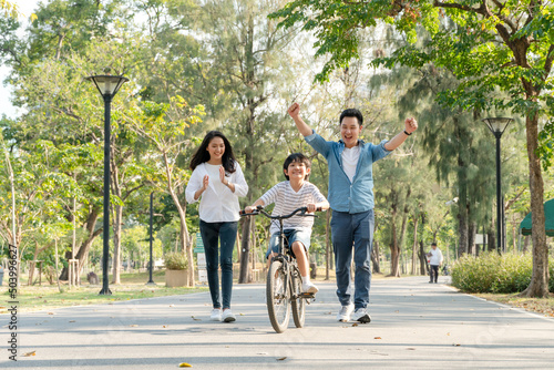 Happy Asian father and mother teach their son to ride a bicycle, Cheerful parent raise hands up in the air to support kid encouragement, family do activity together at park concept © winnievinzence
