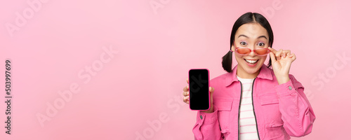 Portrait of asian girl showing mobile phone screen, reacting surprised, standing over pink background