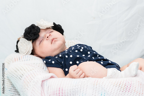 beautiful latina baby girl resting on a white sheet, dressed in a blue romper suit, white stockings and a black and white flower headband. maternity concept photo