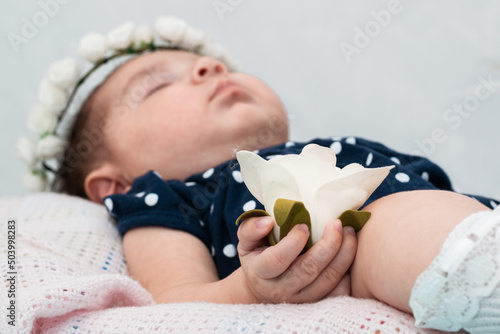 close-up detail of an artificial white rose held by the tender little hand of a newborn baby girl lying in her cradle, dressed in a dark blue jumpsuit and white stockings. photo