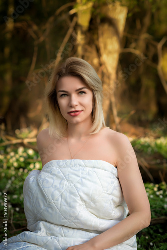 Portrait of a young beautiful fair-haired girl in a light blanket in a forest clearing.