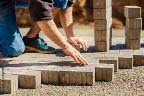 Young man laying gray concrete paving slabs in house courtyard on gravel foundation base. Master lays paving stones. Garden brick pathway paving by professional paver worker. Repairing sidewalk.