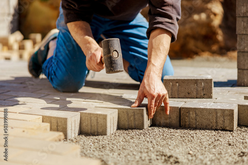 Young man laying gray concrete paving slabs in house courtyard on gravel foundation base. Master lays paving stones. Garden brick pathway paving by professional paver worker. Repairing sidewalk.
