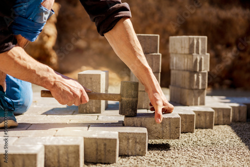 Young man laying gray concrete paving slabs in house courtyard on gravel foundation base. Master lays paving stones. Garden brick pathway paving by professional paver worker. Repairing sidewalk. photo