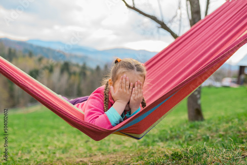 Cute little blond caucasian girl relaxing and having fun in multicolored hammock in outdoor playground. Summer active leisure for kids. Child swinging on hammock. Activities for children