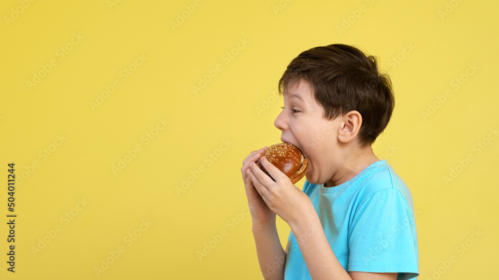 The boy is holding a burger in his hands. Portrait of a funny boy in a blue t-shirt on a yellow background who eats a burger. copy space. concept of unhealthy food. The boy is holding a burger in his hands. Portrait of a funny boy in a blue t-shirt on a yellow background who eats a burger. copy space. concept of unhealthy food.