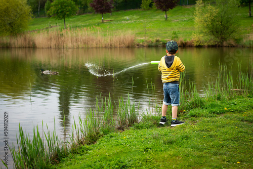 A little cheerful boy shoots water from a play water pump in a park with a lake.