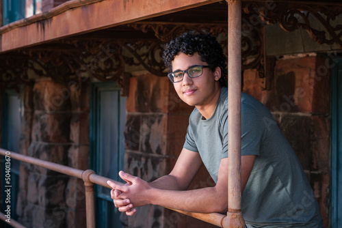 A young man with curly hair leans against an old rusty railing under an awning.