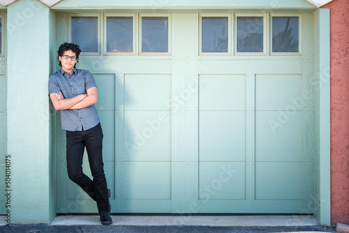 A young man leans against a post in front of a green garage door.