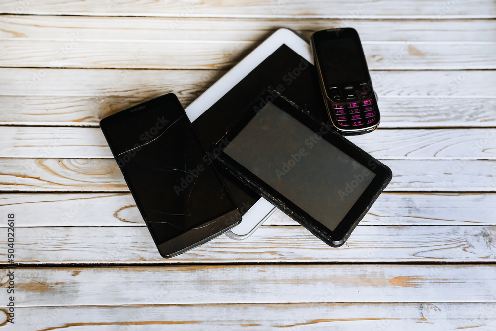 Old, broken, unnecessary, discarded black and white gadgets, mobile smartphones, tablets, electronic devices lie on a wooden background. Old, broken, unnecessary, discarded black and white gadgets, mobile smartphones, tablets, electronic devices lie on a wooden background.