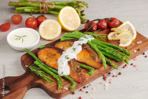 Delicious seasonal green asparagus and sliced smoked salmon on rustic plate with lemon, salt,pepper, of lemon on a wooden cutting board. Baked salmon garnished with asparagus and tomatoes with herbs
