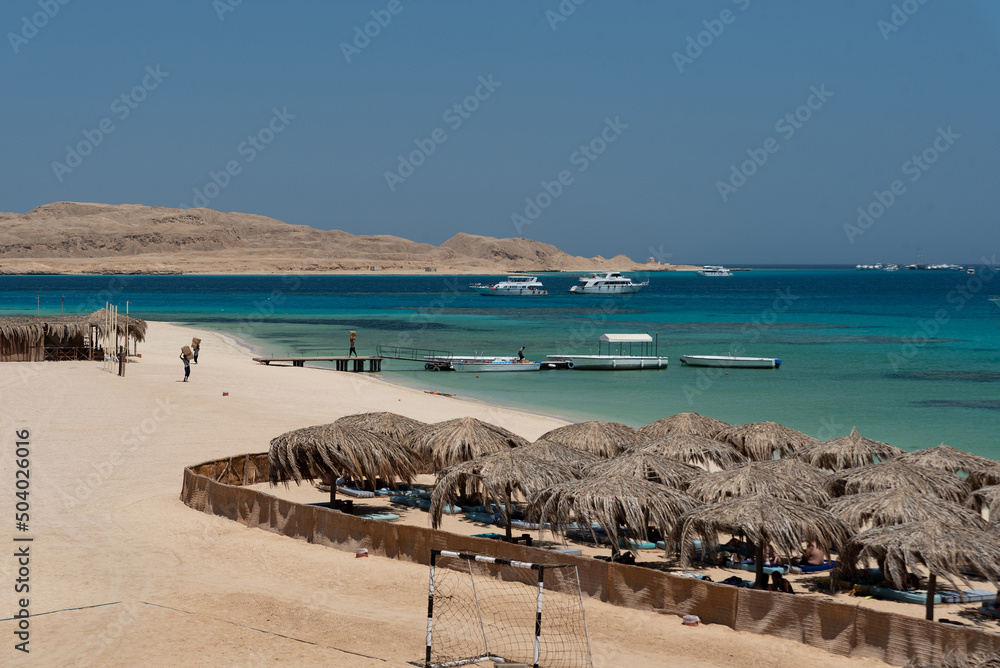 Naklejka premium Holidays in Egypt. Beach with umbrellas made of dry palm leaves, desert and yachts in the blue water of the Red Sea. Vacation and Holidays in Egypt.