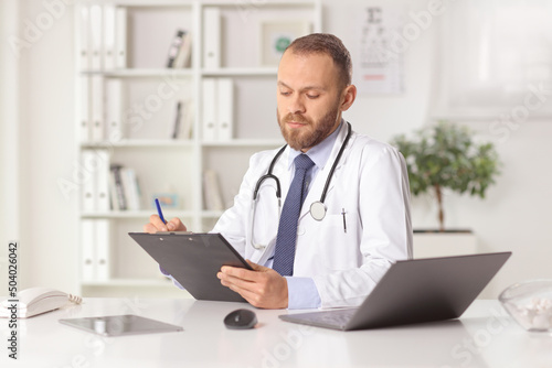 Male doctor in an office sitting and writing a document with a laptop computer on the desk