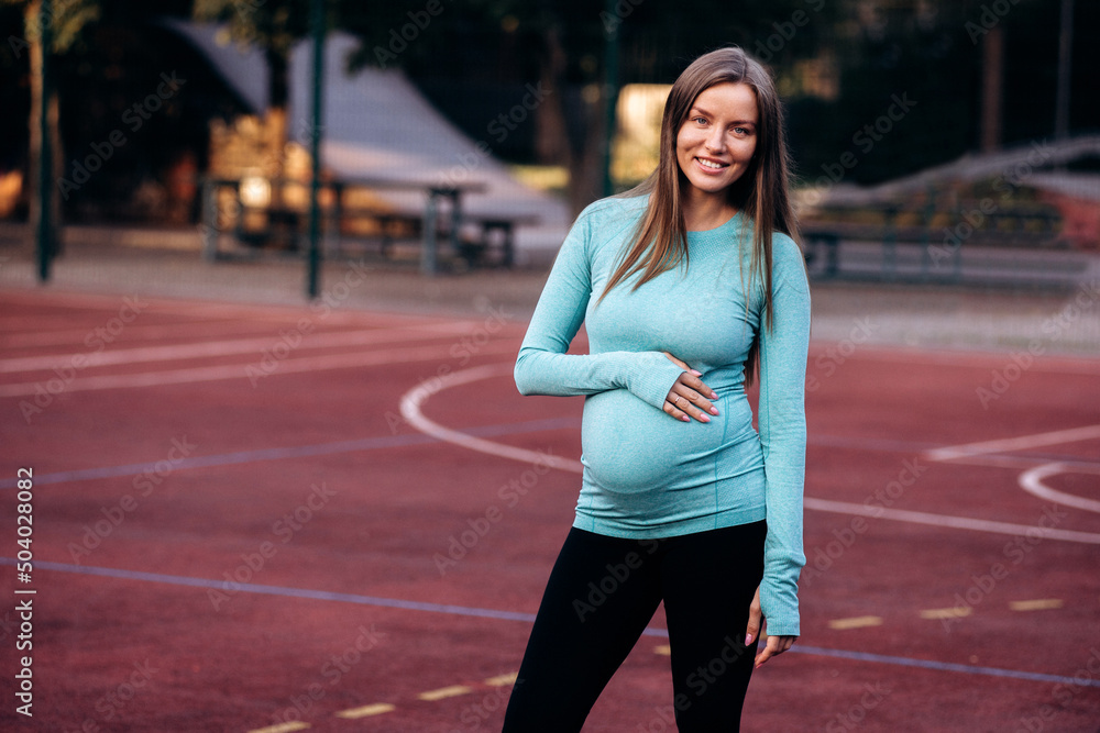 Healthy pregnant woman in sportswear keeps her hands on her stomach, on the sports ground Healthy pregnant woman in sportswear keeps her hands on her stomach, on the sports ground