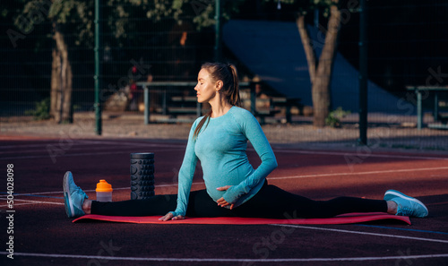 Pregnant performs stretching and sits on the twine on yoga mat on sportsground. Morning exersises of pregnant woman photo