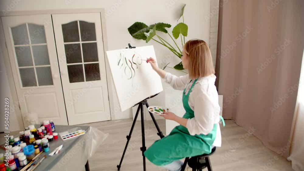Woman in an apron drawers with brush on canvas, there are jars of paint and different brushes at table, side view. Woman draws in living room on an easel. Hobby concept Woman in an apron drawers with brush on canvas, there are jars of paint and different brushes at table, side view. Woman draws in living room on an easel. Hobby concept