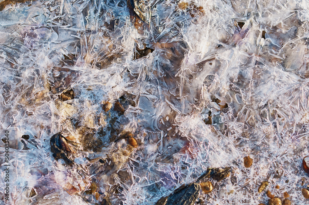 Thin ice in the morning on a rocky beach. Sunrise. Night frosts. Close view. Natural abstract texture. Changeable weather in spring.