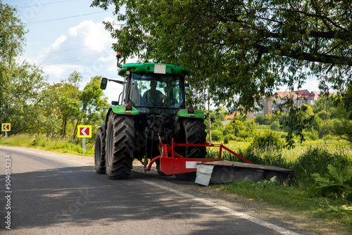 Big modern industrial tractor machine cutting green grass with mowing equipment along country roadside. Road lawn mower machinery vehicle highway maintenance service outdoors on sunny day photo