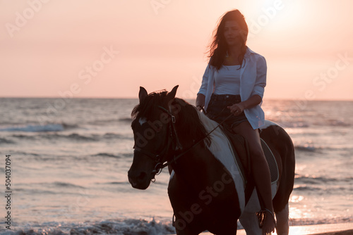 Woman in summer clothes enjoys riding a horse on a beautiful sandy beach at sunset. Selective focus 