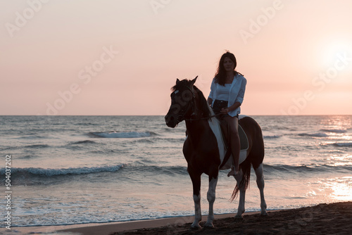 Woman in summer clothes enjoys riding a horse on a beautiful sandy beach at sunset. Selective focus 