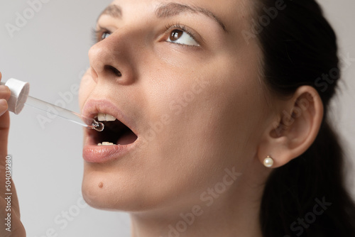 Portrait of dark haired young woman taking oil supplement with glass dropper. CBD hemp medicine oil applying on the tongue  photo