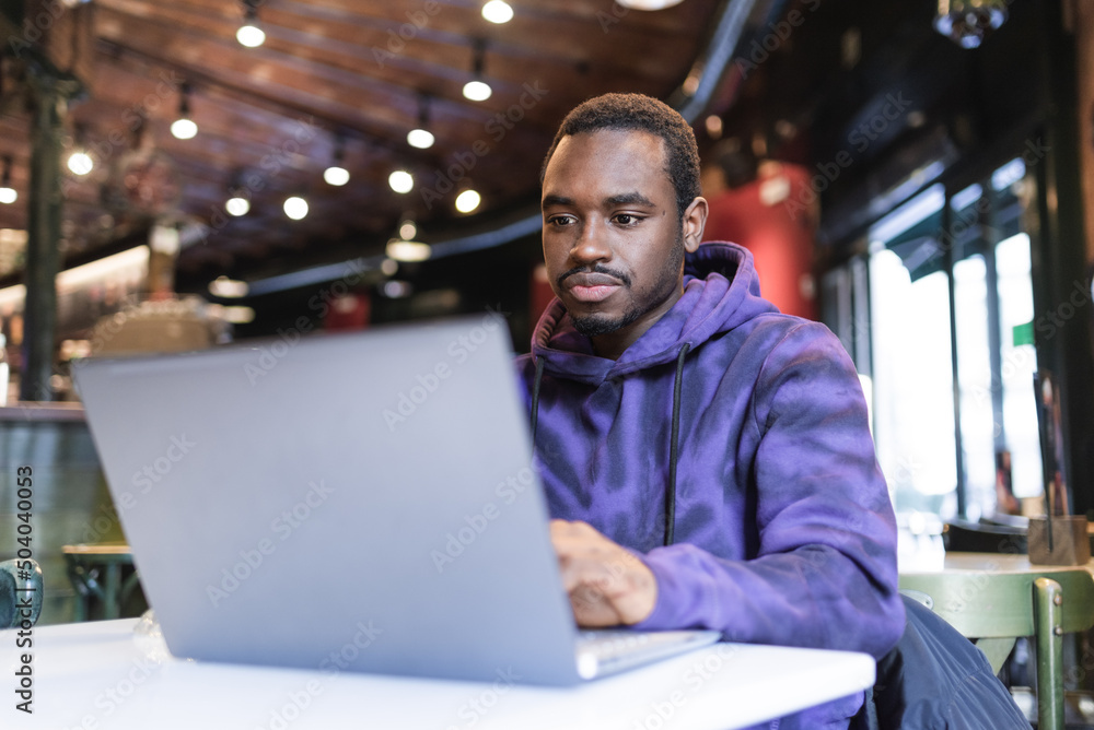 Concentrated African American male freelancer wearing casual hoodie sitting at table and typing on netbook while working on online project in cafe Concentrated African American male freelancer wearing casual hoodie sitting at table and typing on netbook while working on online project in cafe