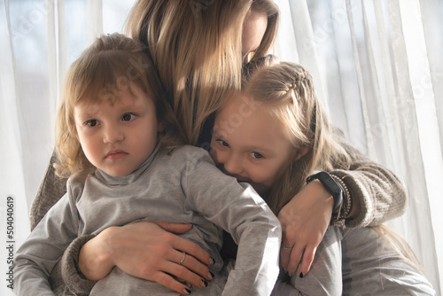 Portrait of a beautiful young woman with two little daughters at home against the background of the window. Family spend leisure time in cozy home environment