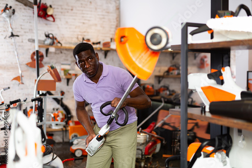 African-american man selecting strimmer in salesroom of gardening tools shop.