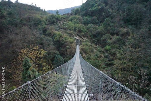 Fototapeta Naklejka Na Ścianę i Meble -  Hanging steel bridge to Nepali village across the natural landscape of rocky stream and green mountain ridge- Nepal