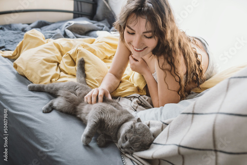 Attractive young woman plays and pets a British shorthair gray cat while lying in bed. Pet and care